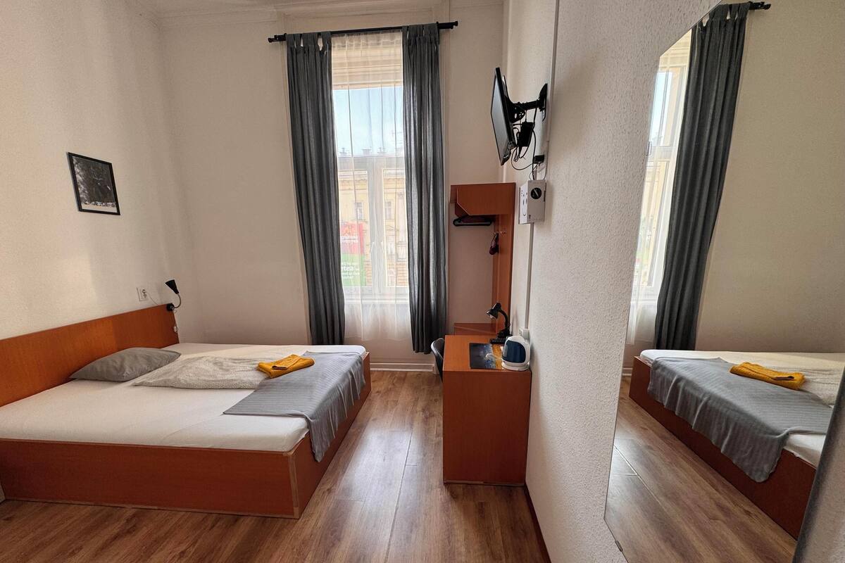 Une chambre avec deux lits, un plancher en bois, un bureau avec une chaise, un miroir et une fenêtre avec des rideaux.