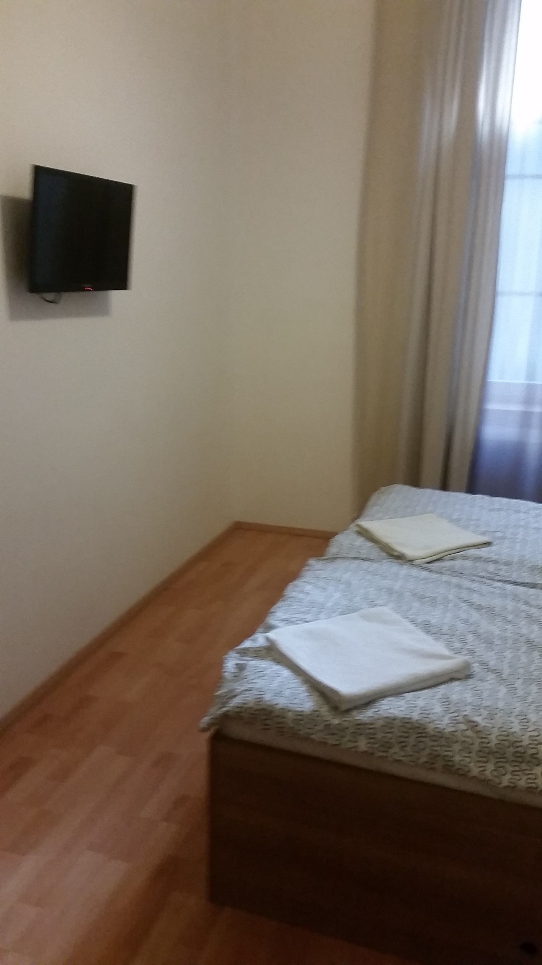 Egy hálószoba, amelyben található egy ágy, egy falra szerelt, síkképernyős televízió és egy ablak, amelyen függöny van.