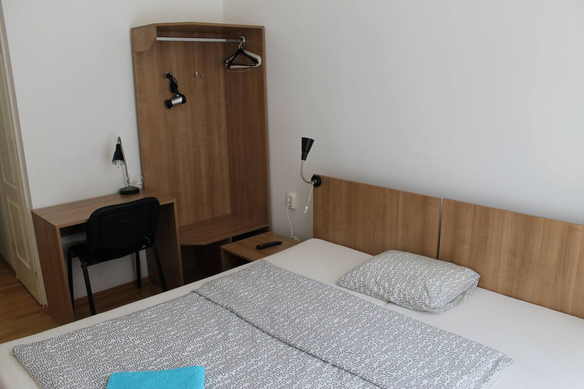 Une chambre d’hôtel avec une armoire en bois, un bureau avec une chaise, un lit avec une literie à motifs et une table de chevet.