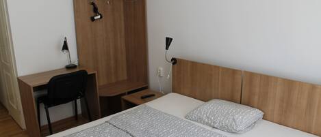 A hotel room with a wooden wardrobe, a desk with a chair, a bed with patterned bedding, and a bedside table.