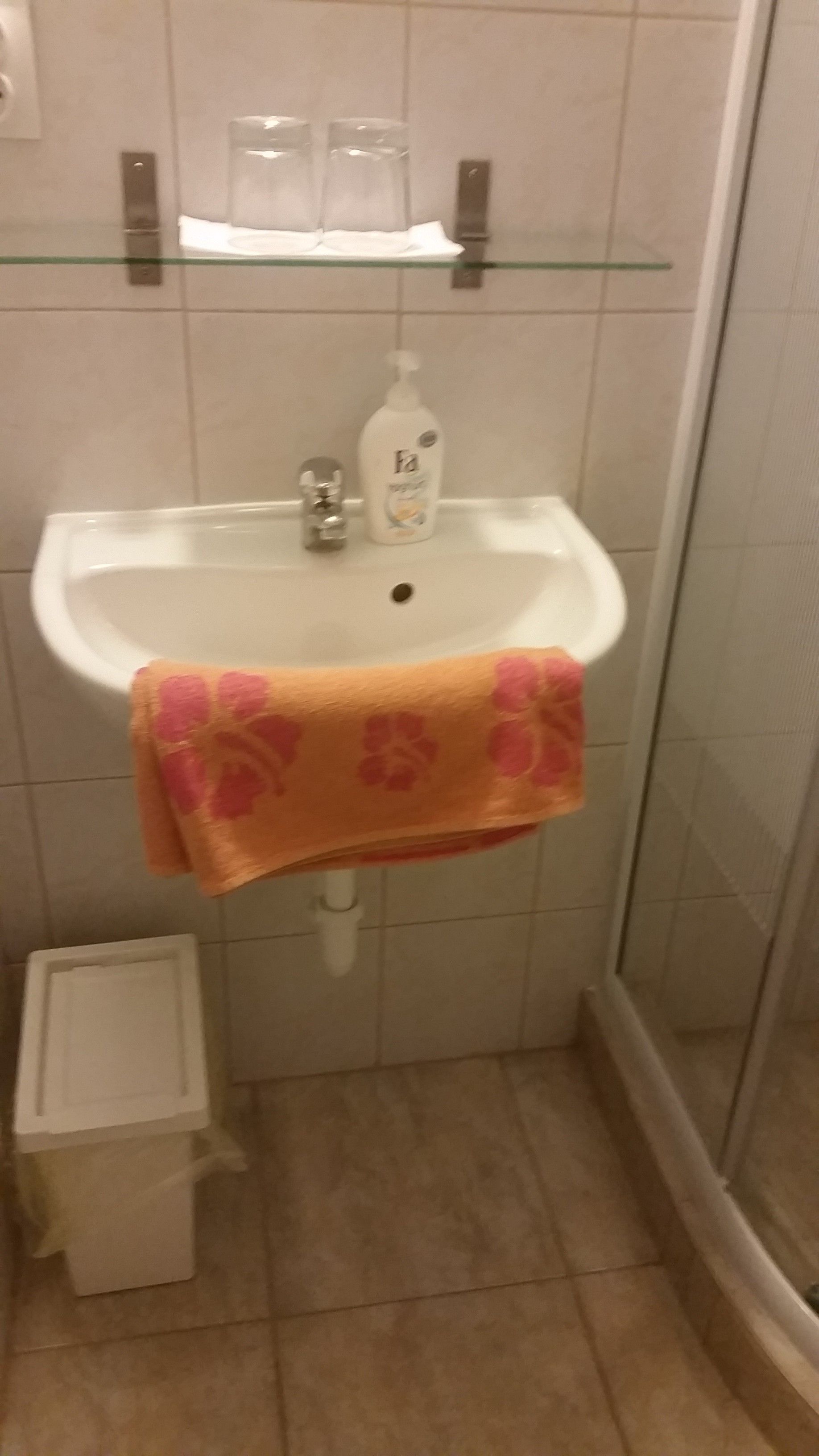 Une salle de bain avec une étagère en verre au-dessus d’un lavabo blanc, un flacon de savon liquide et une serviette à motifs floraux roses suspendue au lavabo.