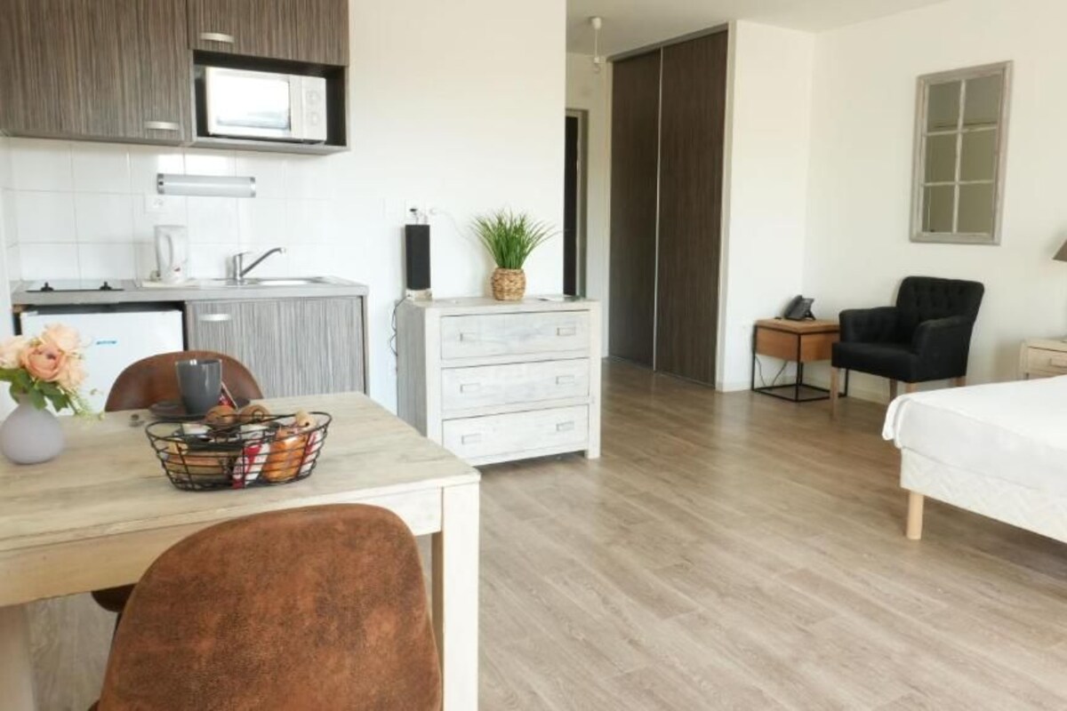 Una habitación de hotel moderna con suelo de madera, una cama, un escritorio con silla, una mesita de noche con lámpara, una silla y un armario.