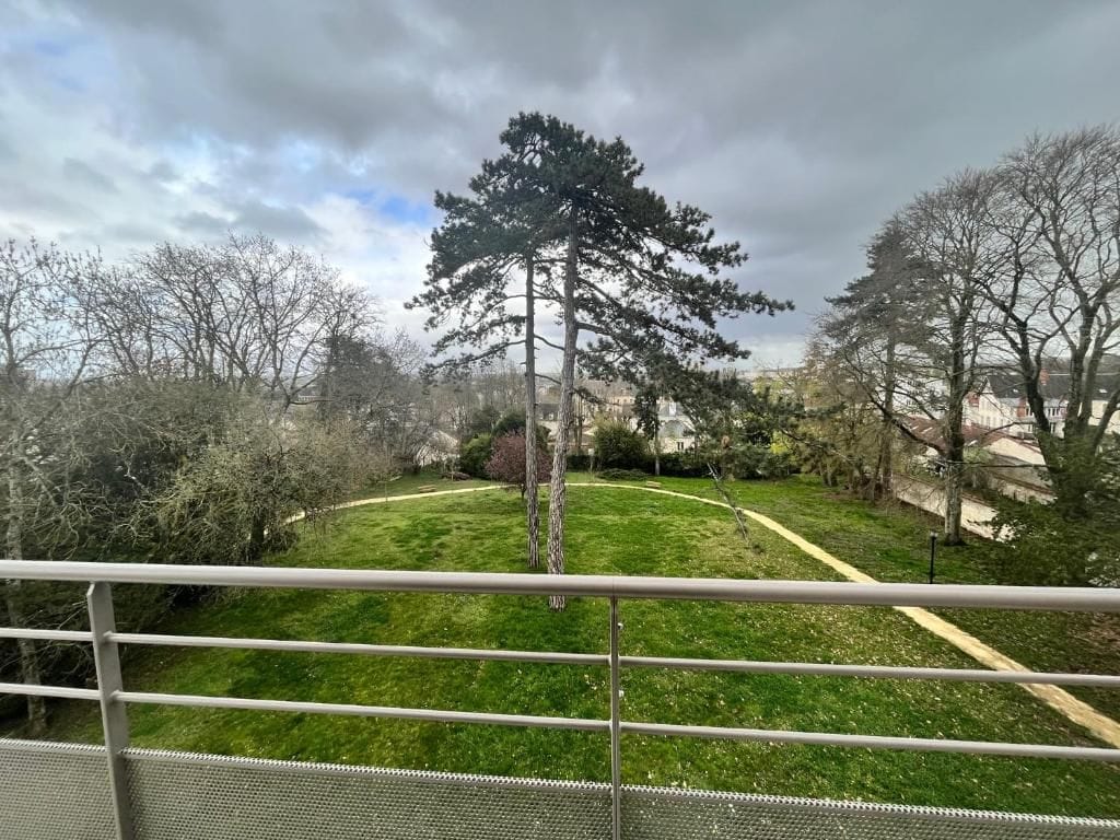 Une vue depuis un balcon doté d’une rambarde en métal, donnant sur un espace verdoyant avec des arbres et un sentier.