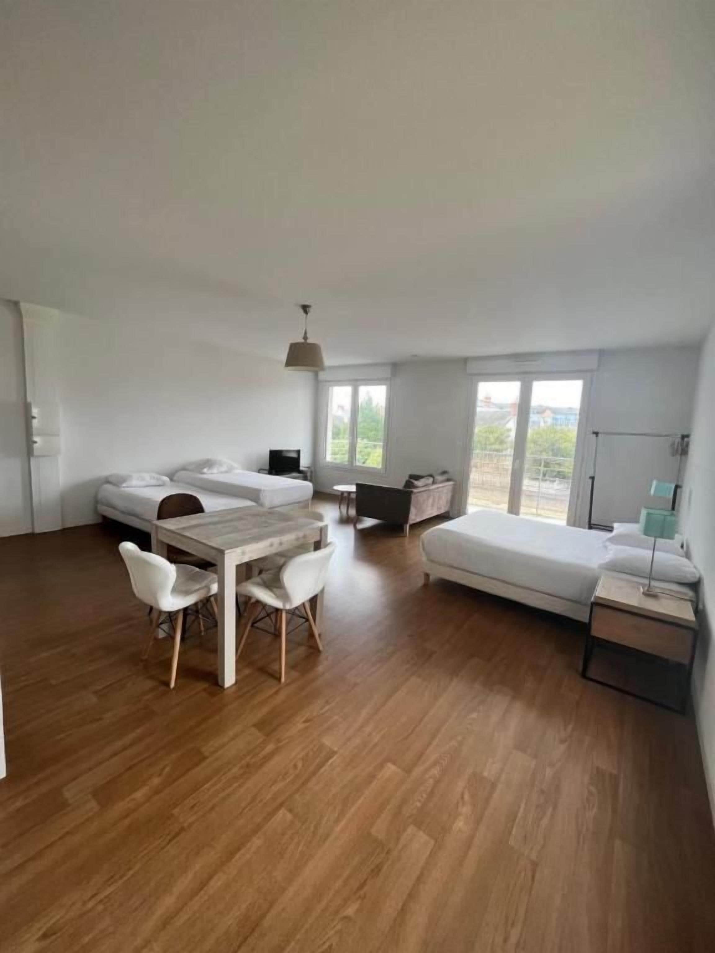 A spacious room with a wooden floor, a bed, a sofa, a dining table with chairs, and a small table with a lamp.
