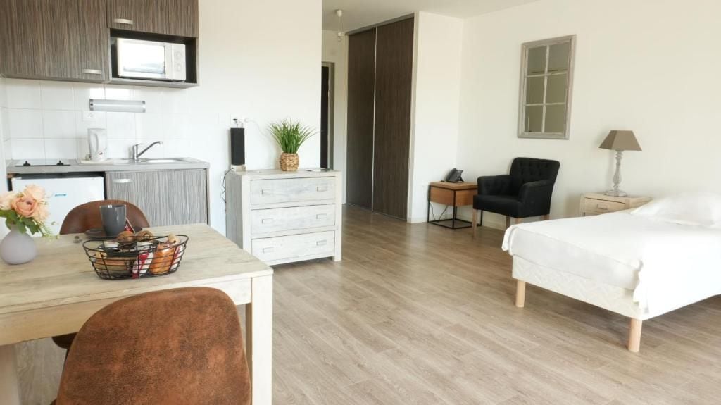 A modern hotel room with a wooden floor, a bed, a desk with a chair, a small table with a lamp, a chair, and a wardrobe.