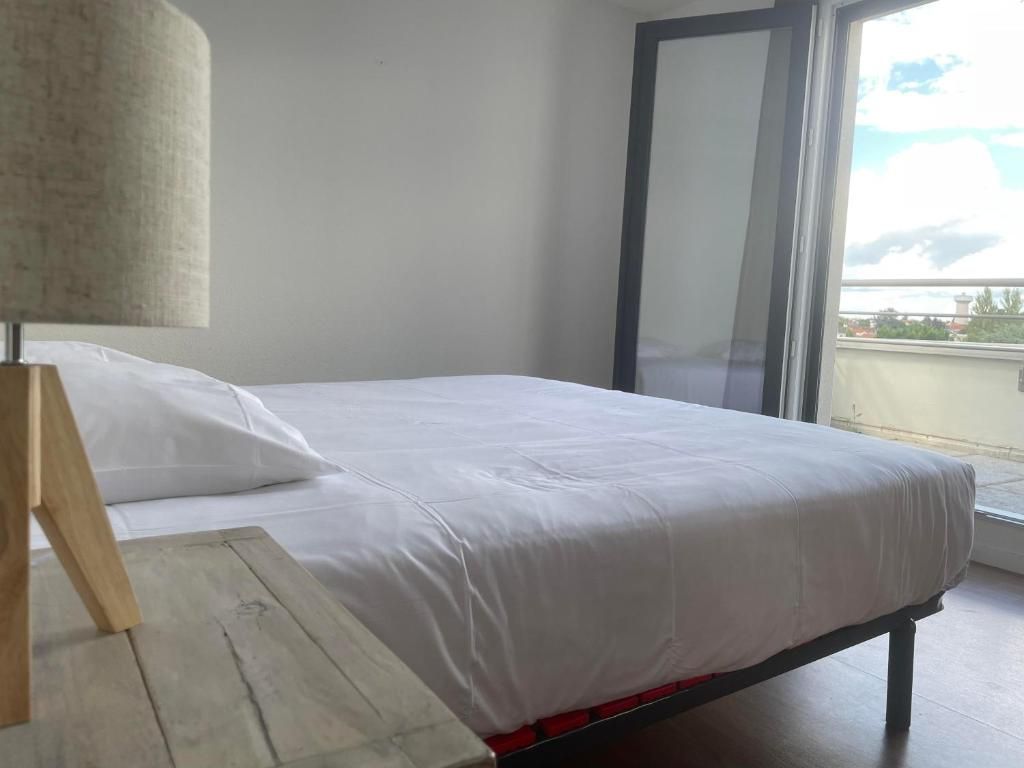 Een bed met witte lakens, een houten nachtkastje en een raam met uitzicht op gebouwen.