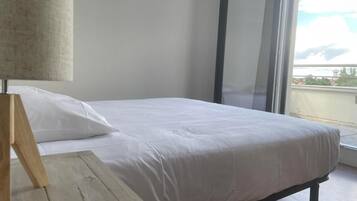 Een bed met witte lakens, een houten nachtkastje en een raam met uitzicht op gebouwen.