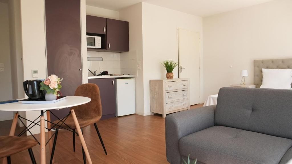 A modern living room with a grey sofa, a dining table with chairs, a kitchen area with cabinets and a microwave, and a small bedside table with a lamp.