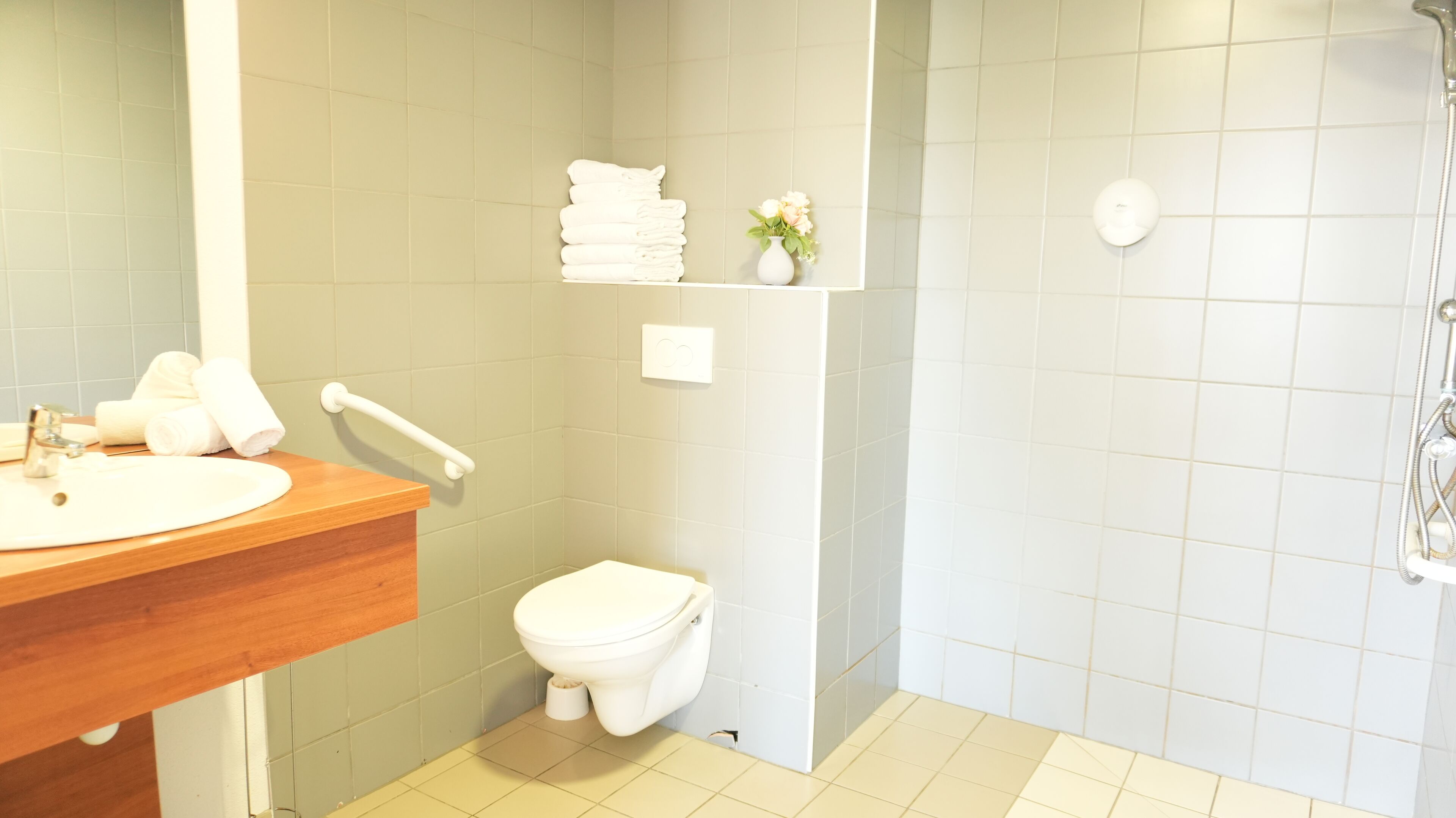 A bathroom with a wooden vanity, a toilet, a shower area, and a shelf with towels and a vase.