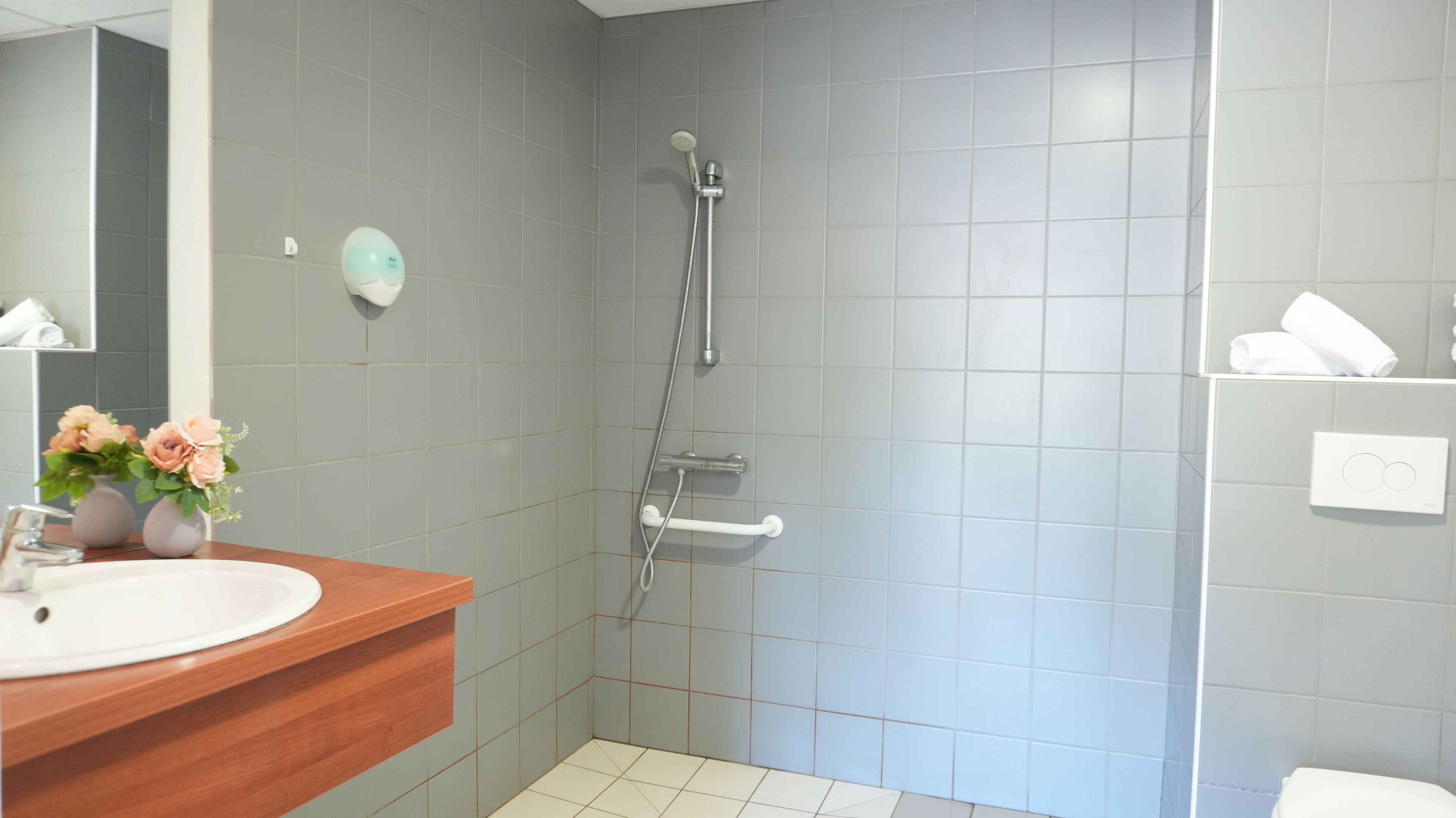 Ein modernes Badezimmer mit einer grauen Duschkabine, einem Holzlagerflächenstück und einem Waschbecken in Weiß sowie einer kleinen Blumenkomposition.