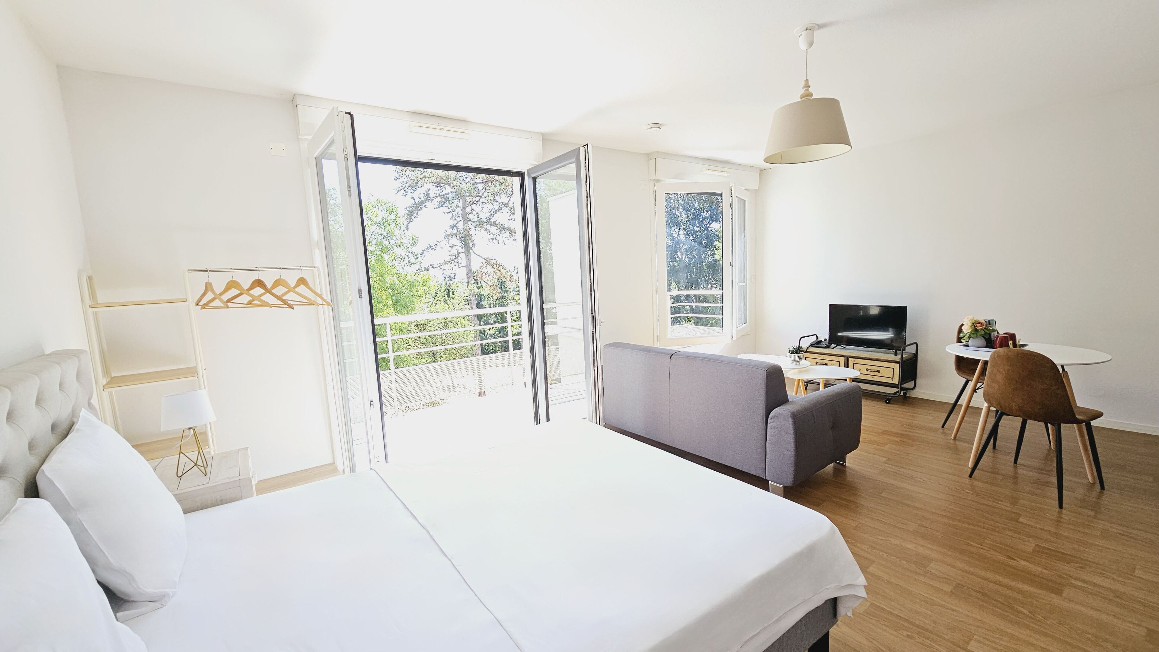 A modern hotel room with a large bed, a grey sofa, a TV, and a balcony with a view of trees.