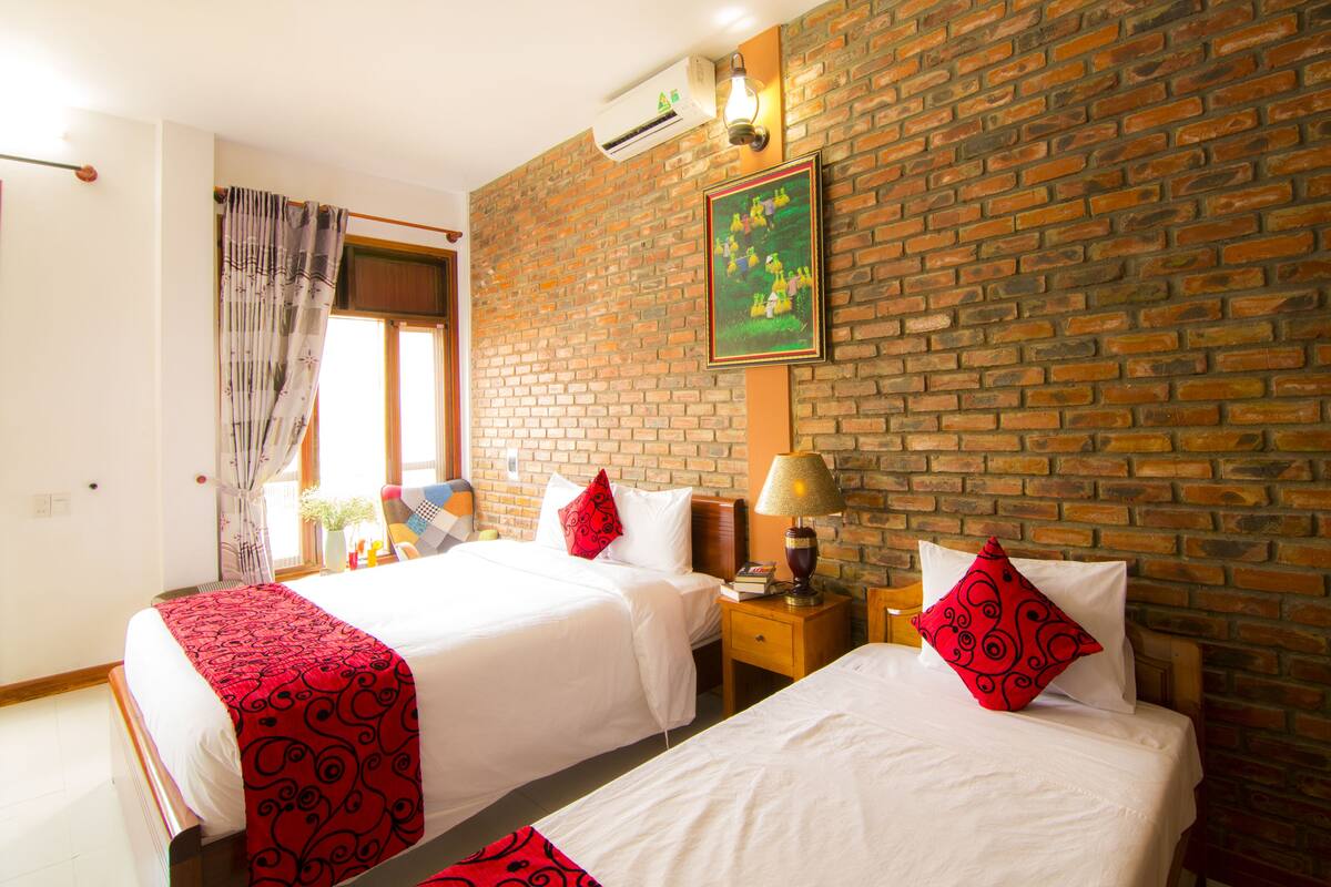 Een hotelkamer met twee bedden, een bakstenen muur, een raam met gordijnen, een lamp en een ingelijste foto.