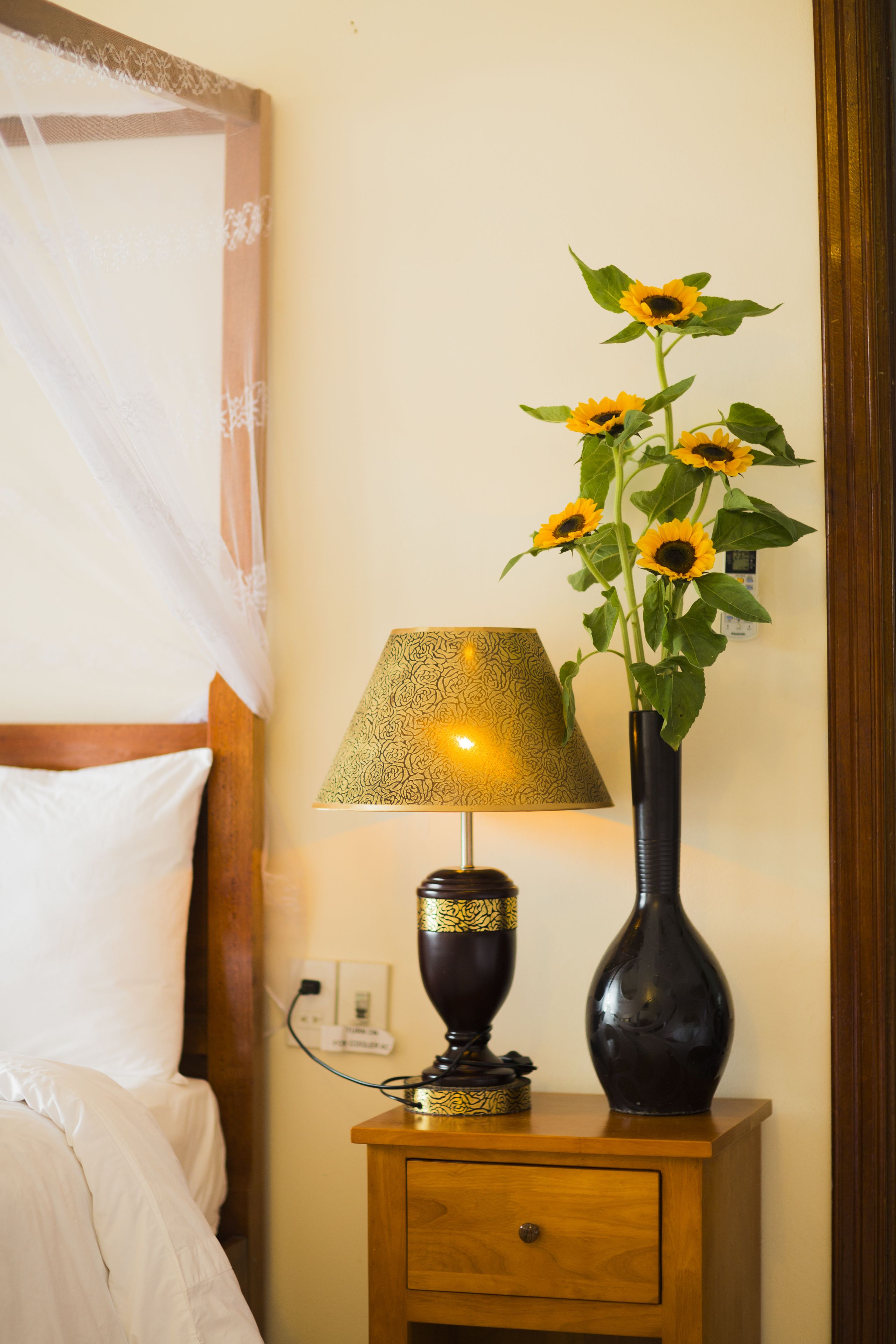 Una cama con ropa de cama blanca, una mesita de noche con lámpara y un florero con girasoles, y un enchufe en la pared.