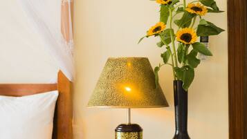 Una cama con ropa de cama blanca, una mesita de noche con lámpara y un florero con girasoles, y un enchufe en la pared.
