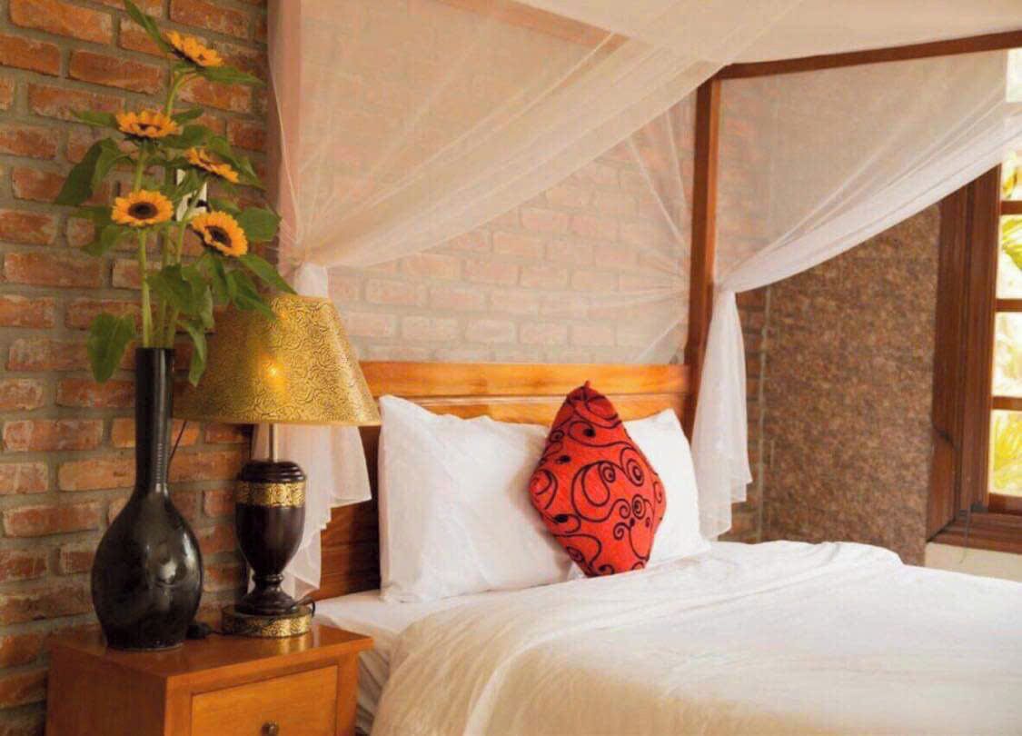 A bedroom with a brick wall, a wooden headboard, a bed with white bedding, a red patterned pillow, a brass lamp, and a vase with sunflowers.