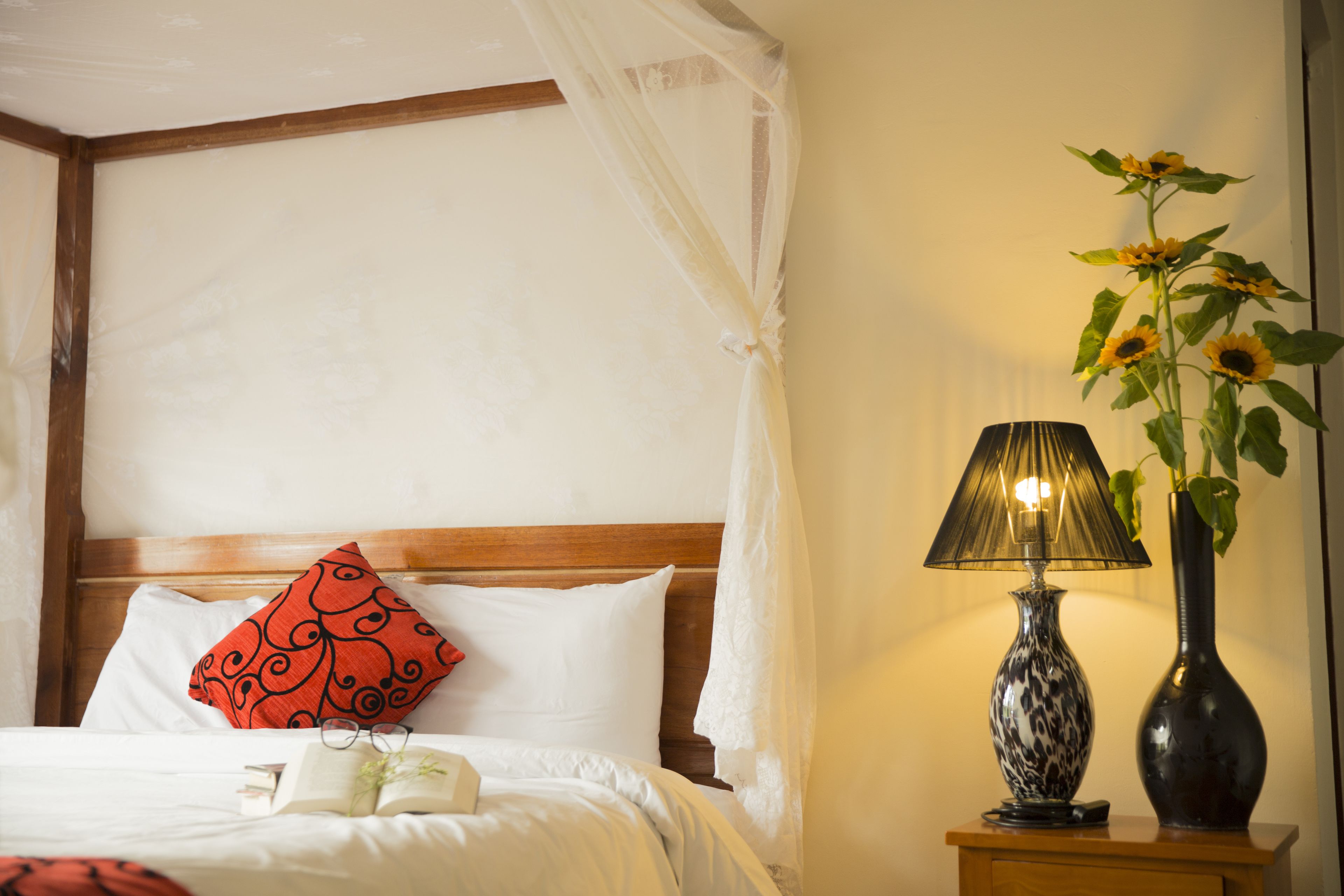 A neatly made bed with a white and red pillow, a bedside table with a lamp and a vase, and a window with a sheer curtain.