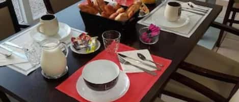 Una mesa de desayuno servida con platos, una jarra de leche, una cesta de croissants, un vaso de jugo y una taza de café.
