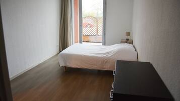 A bedroom with a bed, a wooden dresser, a nightstand with a lamp, and a sliding glass door leading to a balcony.