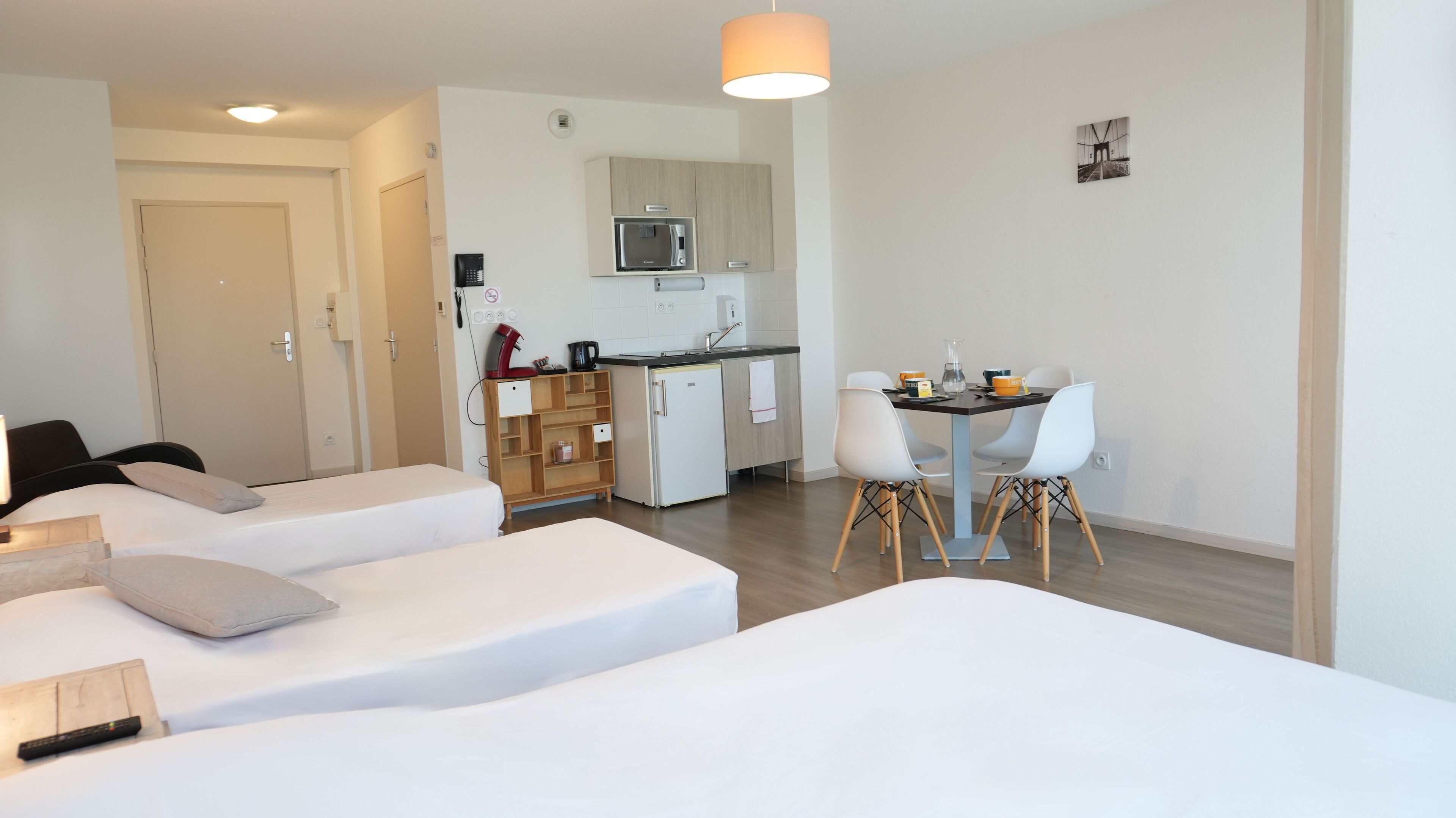 Una habitación de hotel moderna con dos camas individuales, una kitchenette y un comedor con sillas blancas.