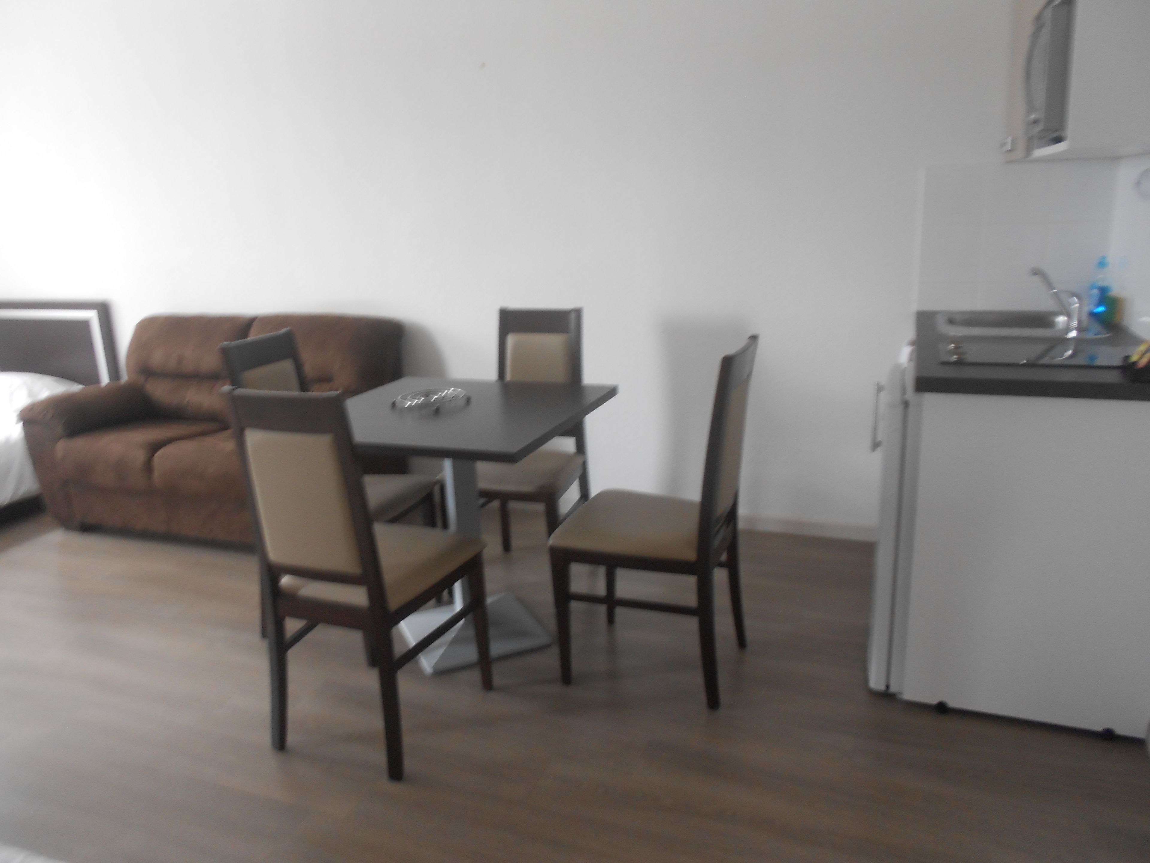 A compact living space with a dining table and four chairs, a small kitchenette, and a brown sofa.