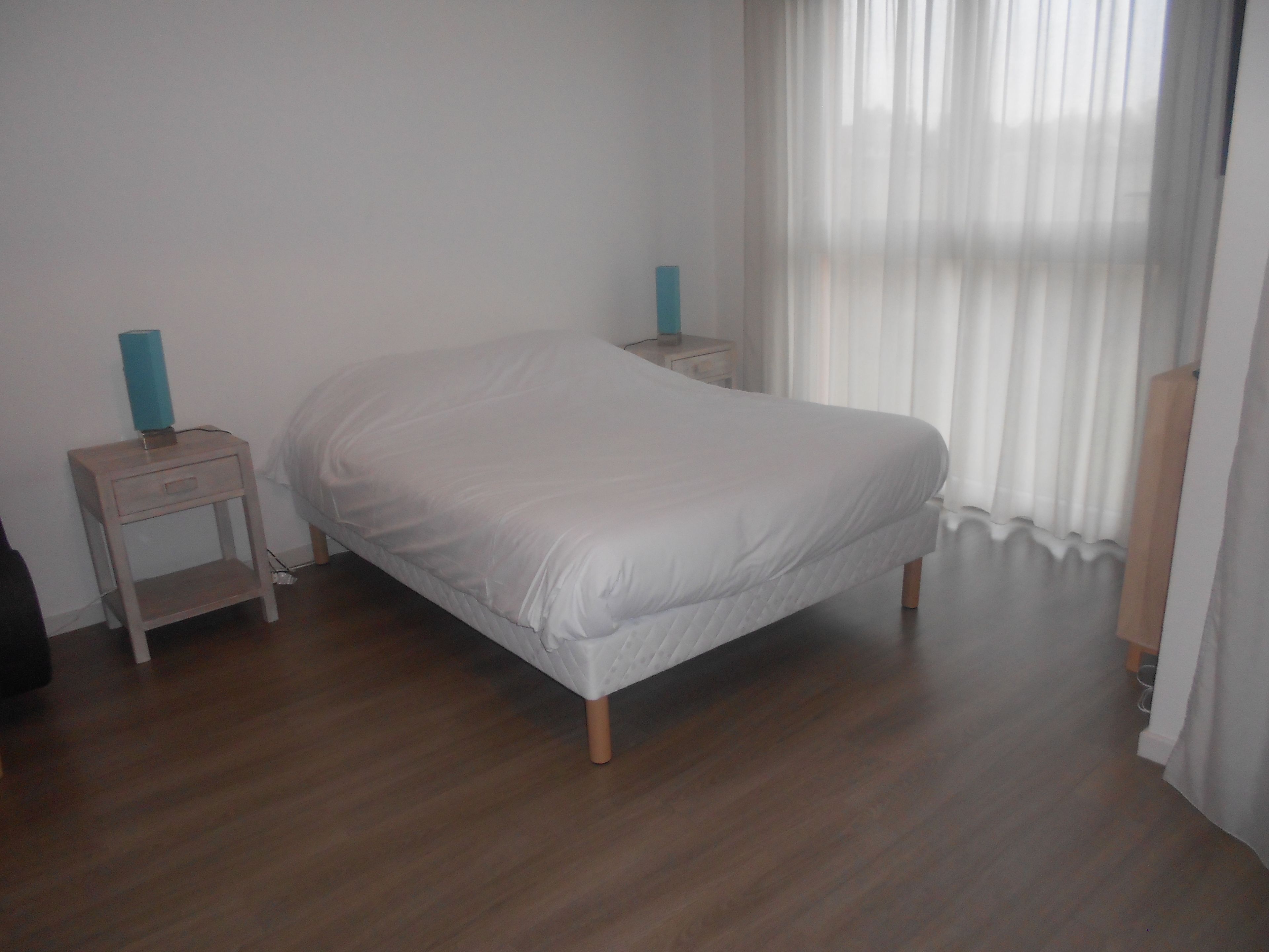 Ein Schlafzimmer mit einem Einzelbett, Holznachttischen und einem Fenster mit durchsichtigen Vorhängen.