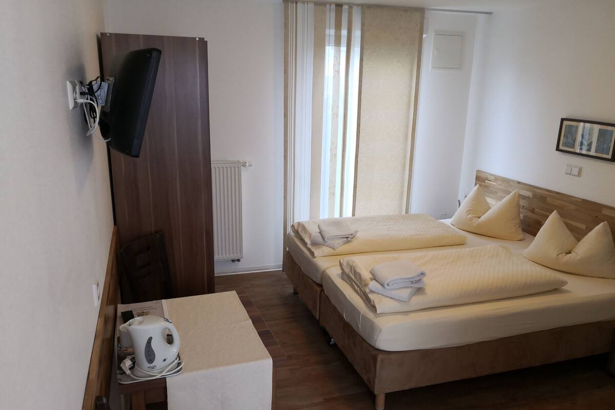 Ein Hotelzimmer mit zwei Betten, einem Schreibtisch mit Wasserkocher, einem an der Wand montierten Fernseher und einem Fenster mit Vorhängen.