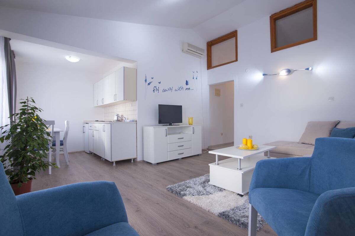A modern living room with a sofa, a coffee table, and a TV. There is a kitchen area with white cabinets and a dining table.