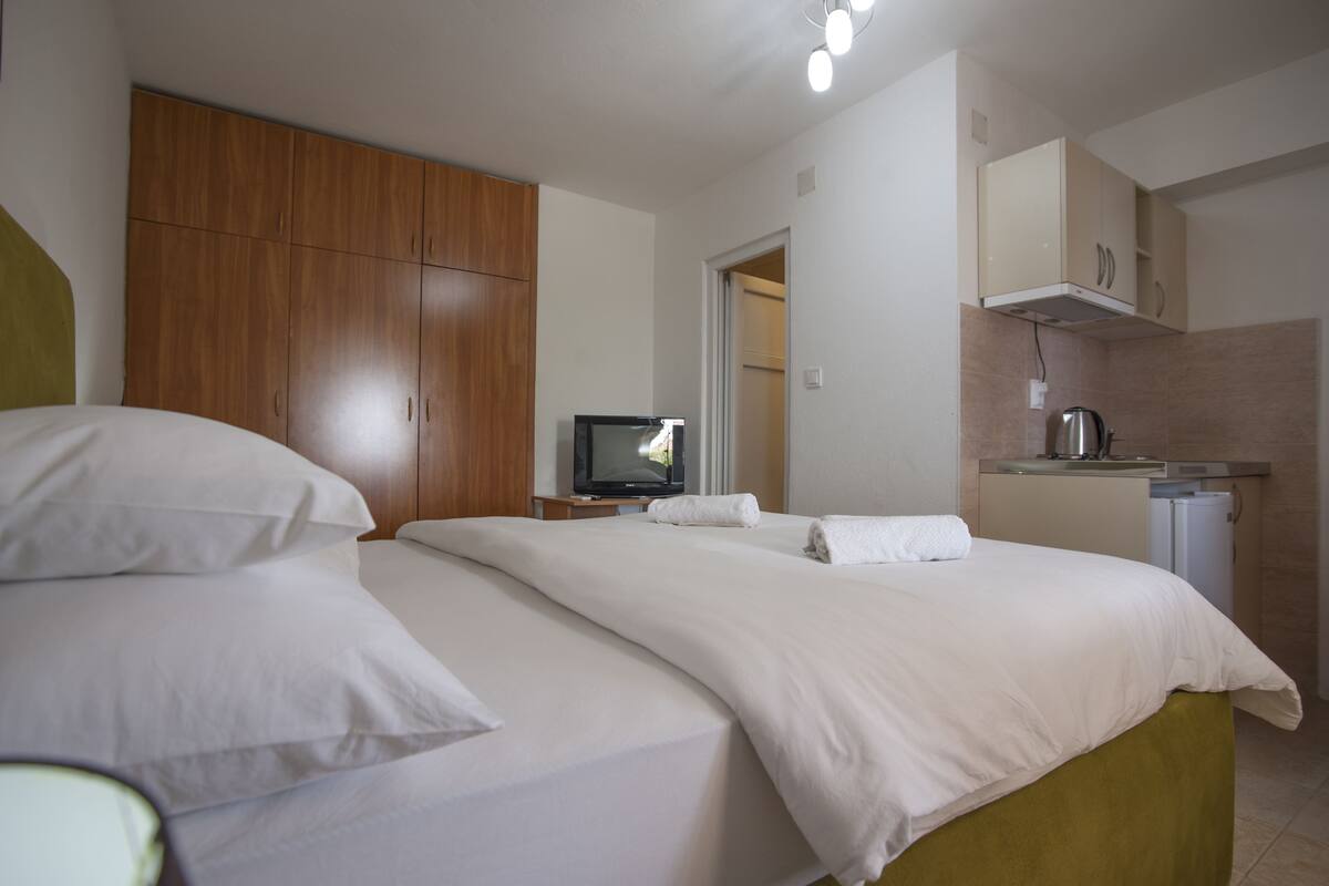 A hotel room with a bed, a television, a small kitchenette, and wooden wardrobe.