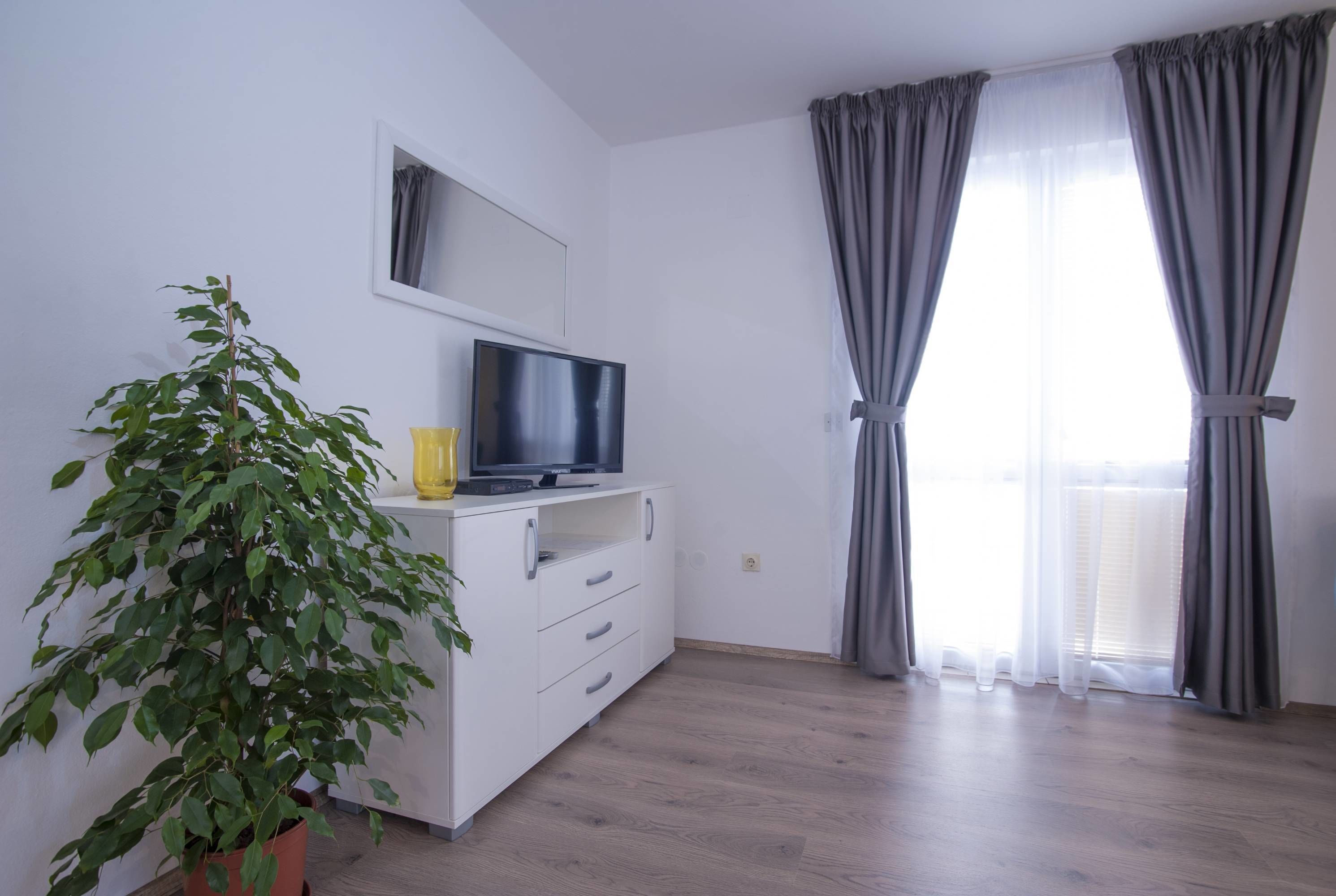 Uma sala de estar moderna com uma televisão de tela plana sobre um armário branco, um vaso de planta e uma grande janela com cortinas cinzas.