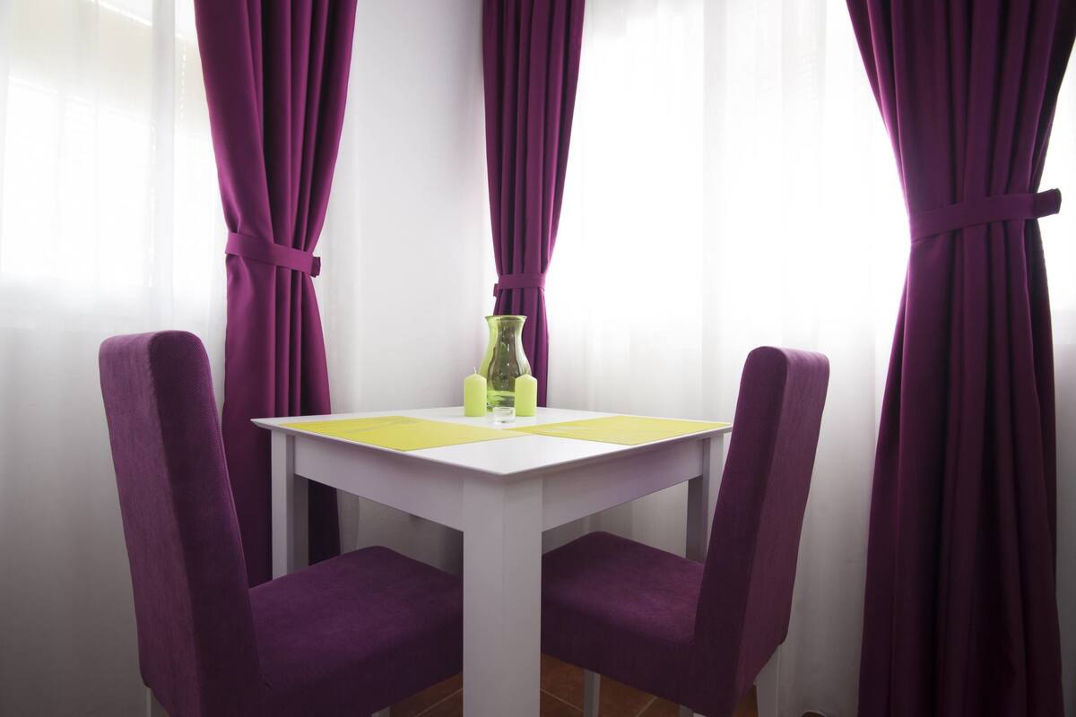 A dining area with a white table and purple chairs, a glass vase with candles, and purple curtains.
