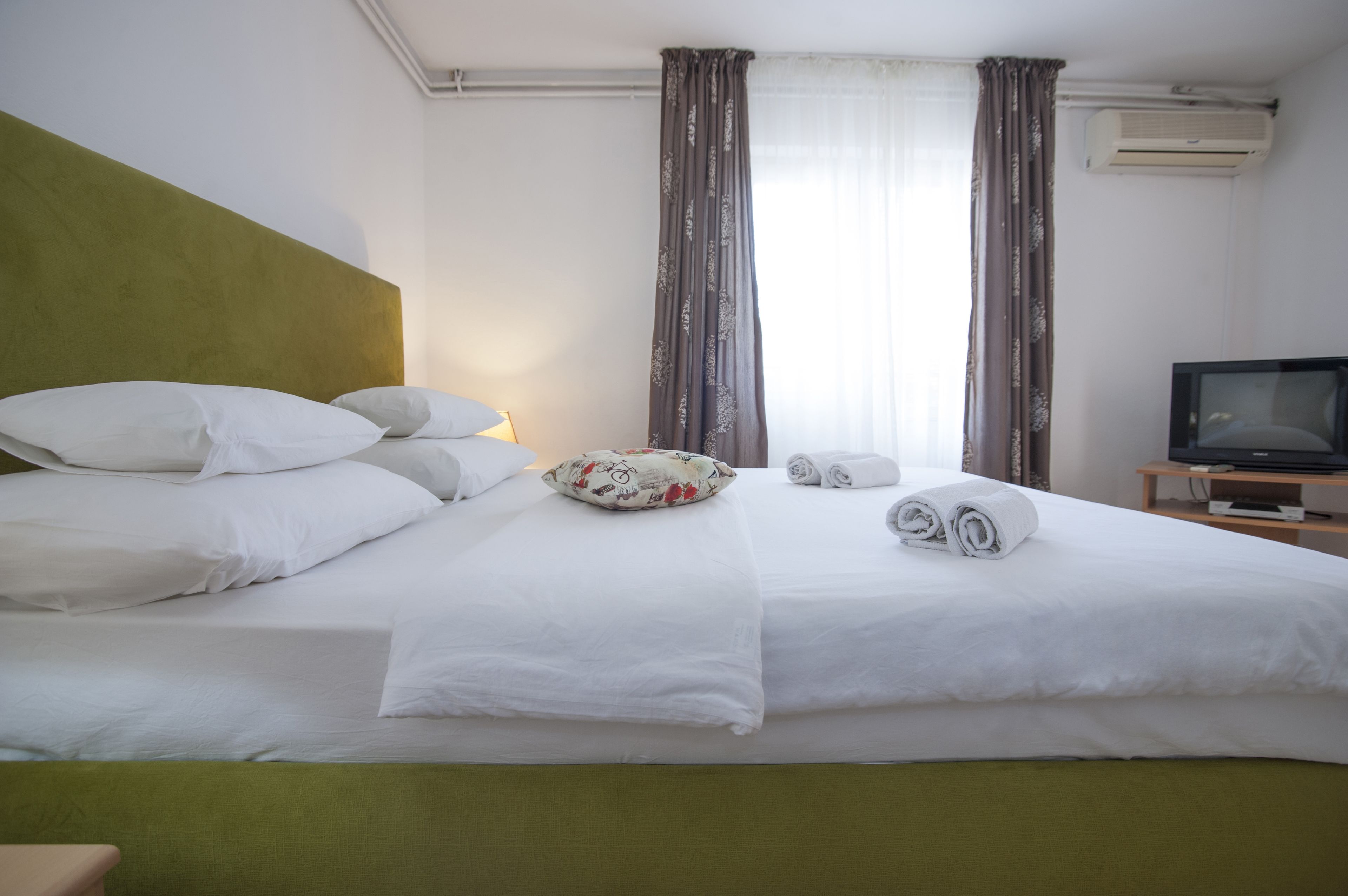 Un lit bien fait, avec des draps blancs, une tête de lit verte et un coussin à motifs floraux. Deux serviettes enroulées sont posées sur le lit. Une télévision est posée sur un meuble en bois, à droite.