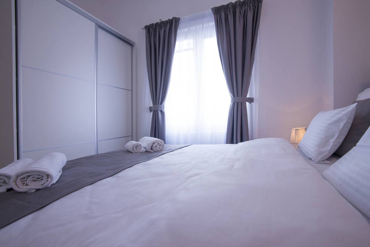 A hotel room with a neatly made bed, two rolled towels, a bedside lamp, and a window with curtains.