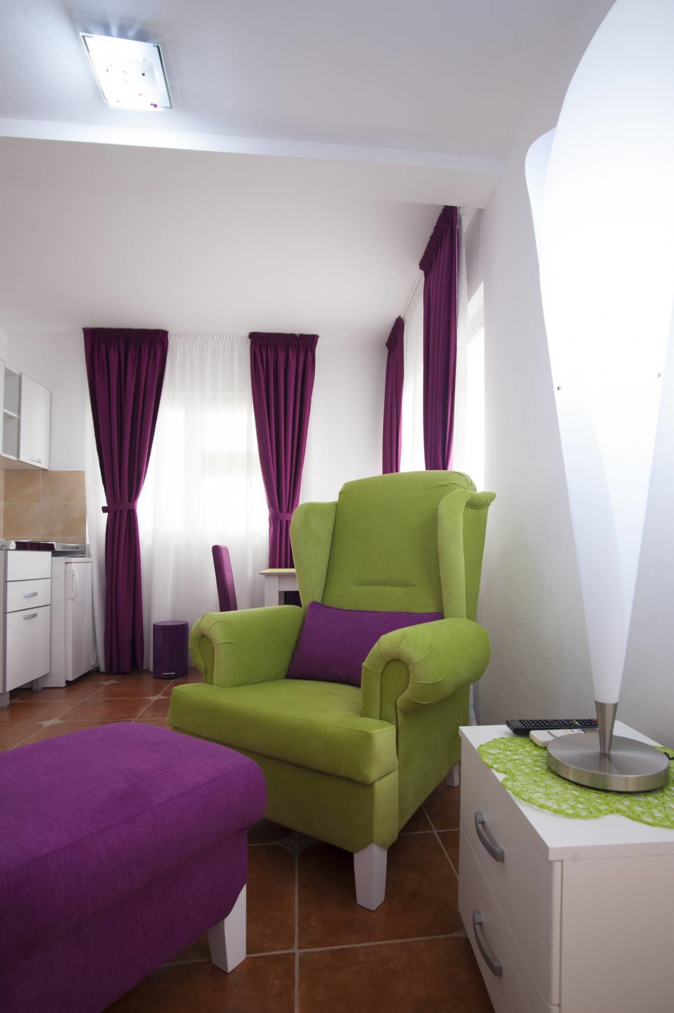 A modern room with a green armchair, a purple ottoman, and a white bedside table with a lamp.