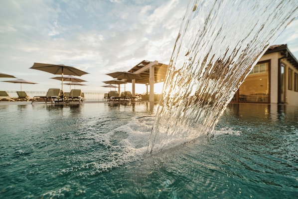 A swimming pool with water cascading over the edge, lounge chairs, umbrellas, and a building in the background.