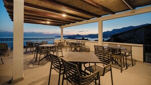Una terraza exterior con mesas y sillas, techo de paja y vista al mar y las montañas.
