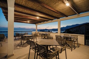 An outdoor terrace with tables and chairs, a thatched roof, and a view of the sea and mountains. - Apartments Grand (Blizikuce)