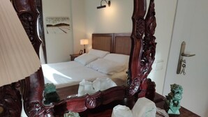 Ein Hotelzimmer mit einem großen, verzierten Spiegel, einem Bett mit weißen Leinentüchern, einem Nachttisch mit Lampe und einer kleinen Statue auf der Kommode.
