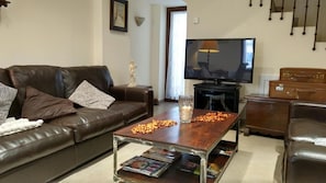 Uma sala de estar com um sofá de couro marrom, uma mesa de centro de madeira com revistas, uma televisão e um armário com malas.