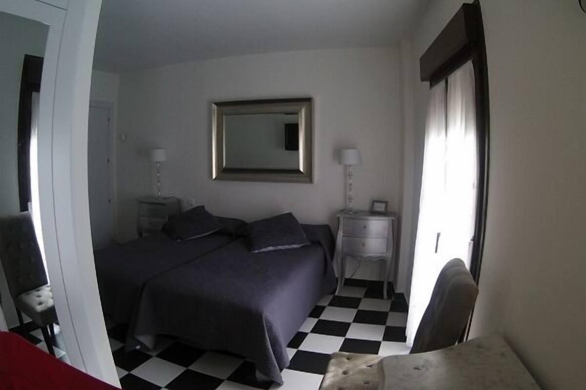 Un dormitorio con piso a cuadros, cama con ropa de cama oscura, dos mesitas de noche con lámparas, un espejo en la pared y una puerta con cortina.