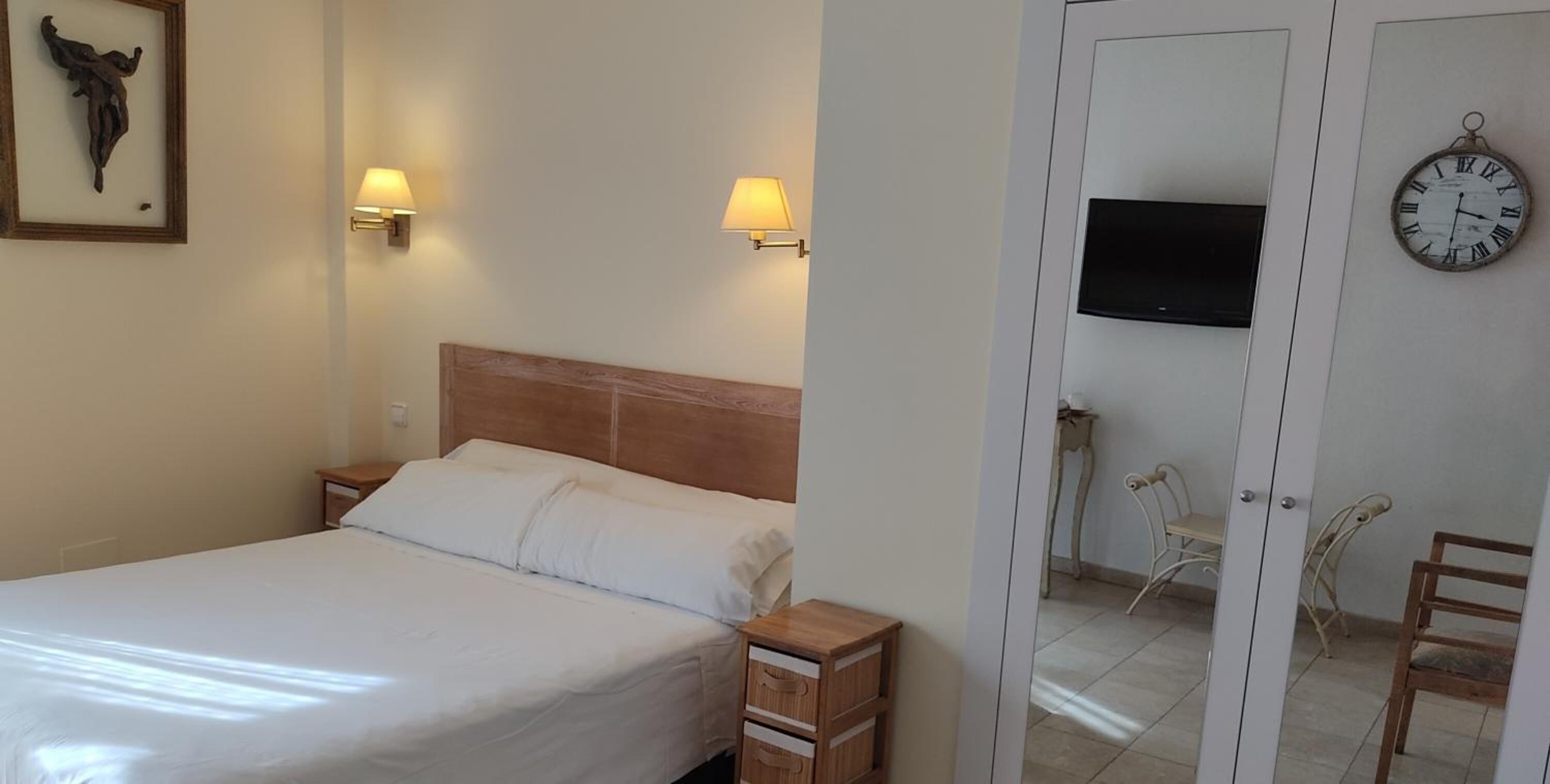 Una cama bien hecha con ropa blanca, dos lámparas fijadas a la pared, una mesita de noche de madera, una obra de arte enmarcada en la pared, un televisor, un reloj y una puerta que da a otra habitación.