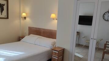 Una cama bien hecha con ropa blanca, dos lámparas fijadas a la pared, una mesita de noche de madera, una obra de arte enmarcada en la pared, un televisor, un reloj y una puerta que da a otra habitación.