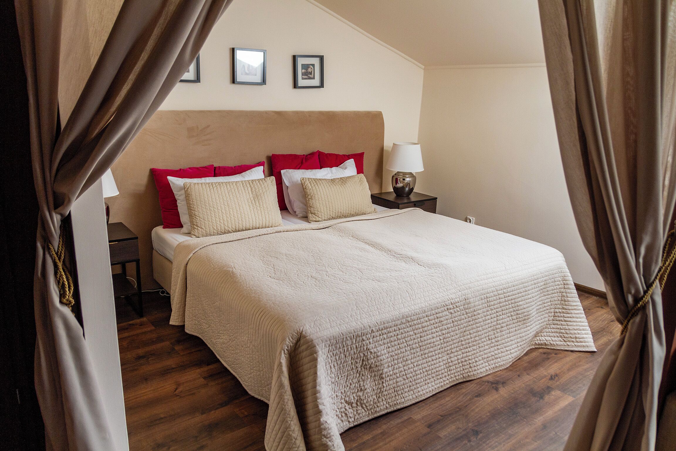 A neatly made bed with a beige quilt, red and white pillows, and a bedside table with a lamp.