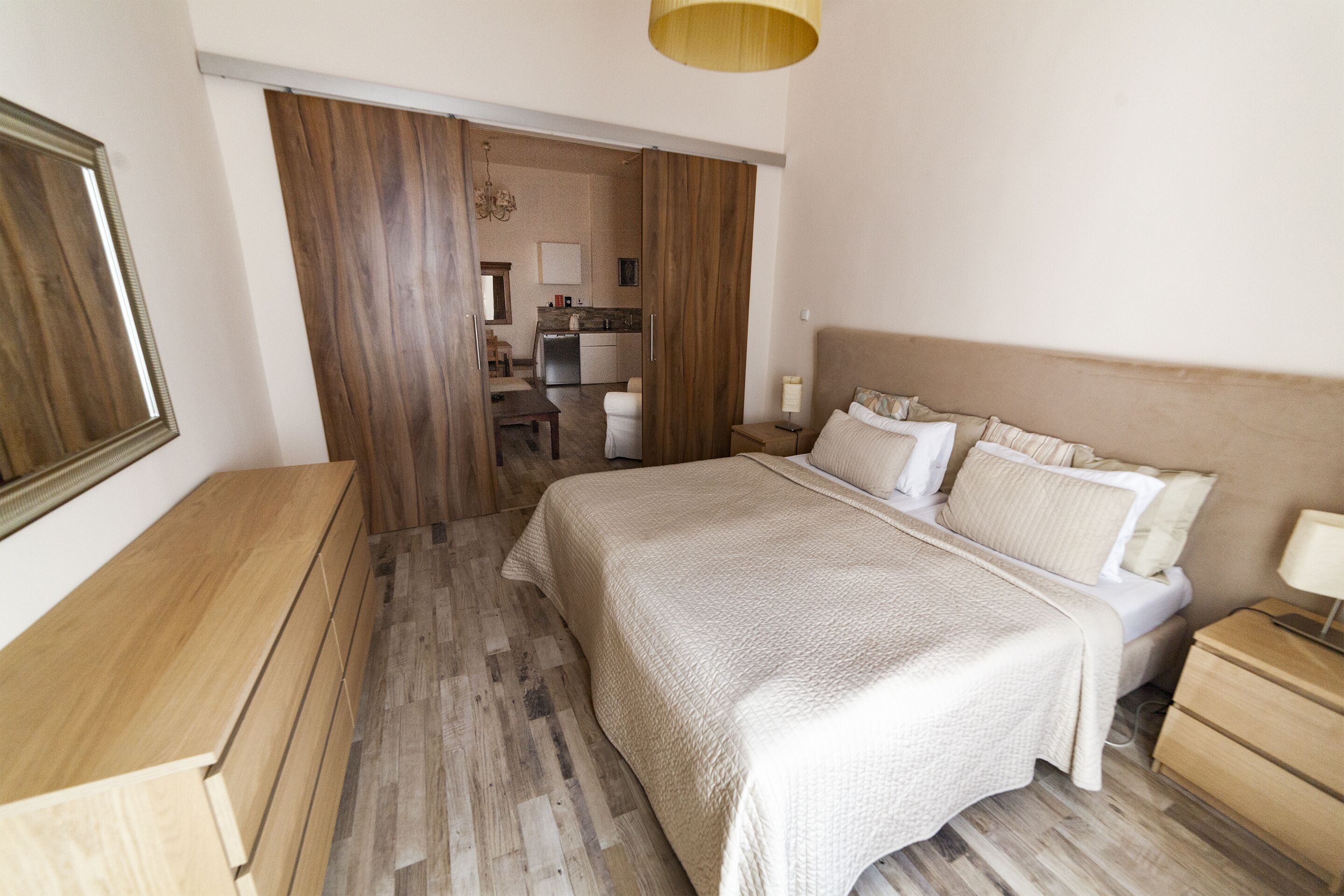 A bedroom with a large bed, wooden floor, a desk, and a kitchenette in the background.