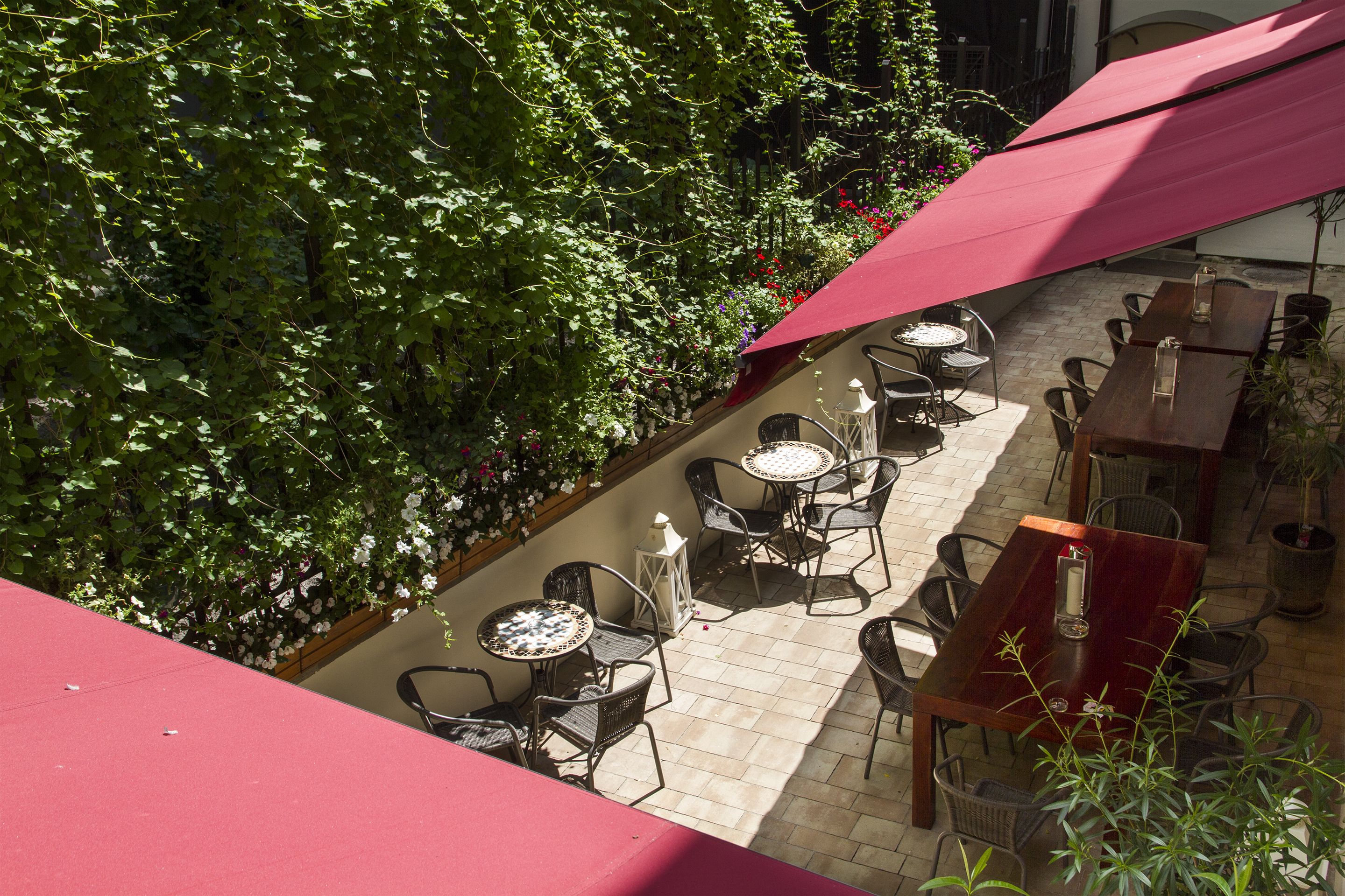 Une terrasse extérieure dotée de plusieurs tables et chaises, entourée d’une végétation luxuriante et protégée du soleil par un auvent rouge.