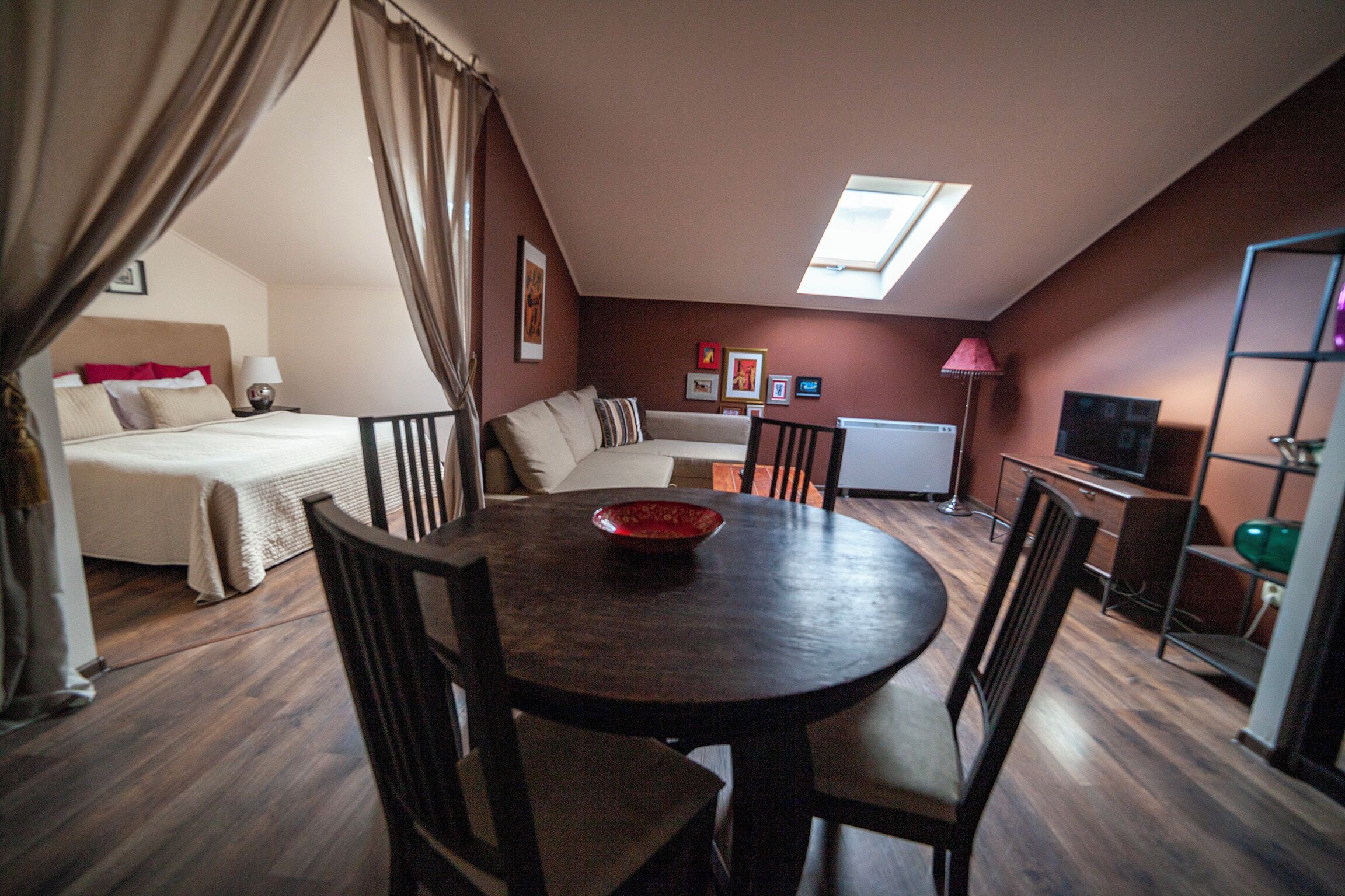 A room with a bed, a sofa, a dining table with chairs, a TV, and a window with curtains.