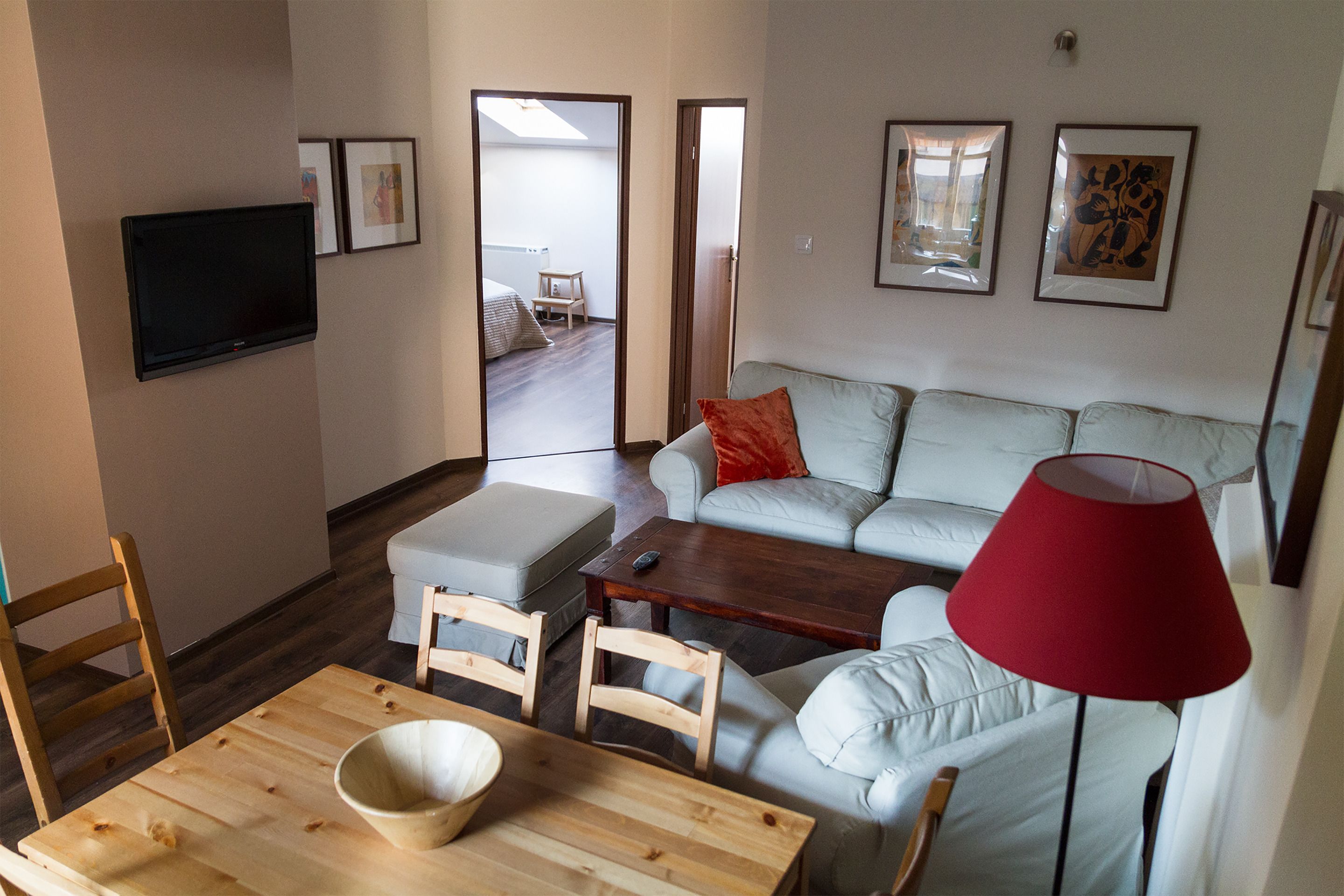 Een moderne woonkamer met een bank, een salontafel en een aan de muur bevestigde televisie. Er hangen ingelijste foto’s aan de muur en staat een rode lamp op de vloer.