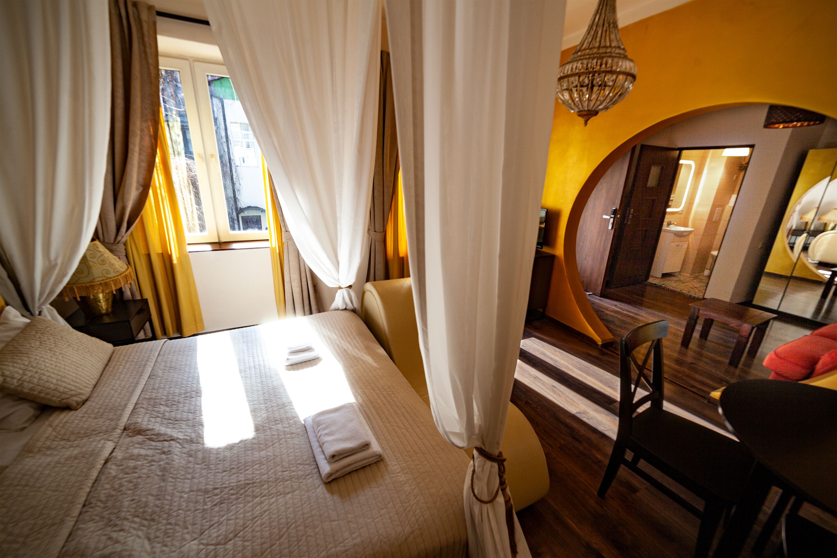 A hotel room with a bed, curtains, a chandelier, a mirror, and a view of the outside.