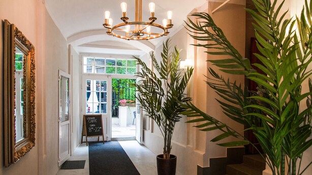 A hallway with a black carpet, a large potted plant, a chandelier, and a sign indicating the lounge coffee is open. - Lounge Apartments (Kraków)