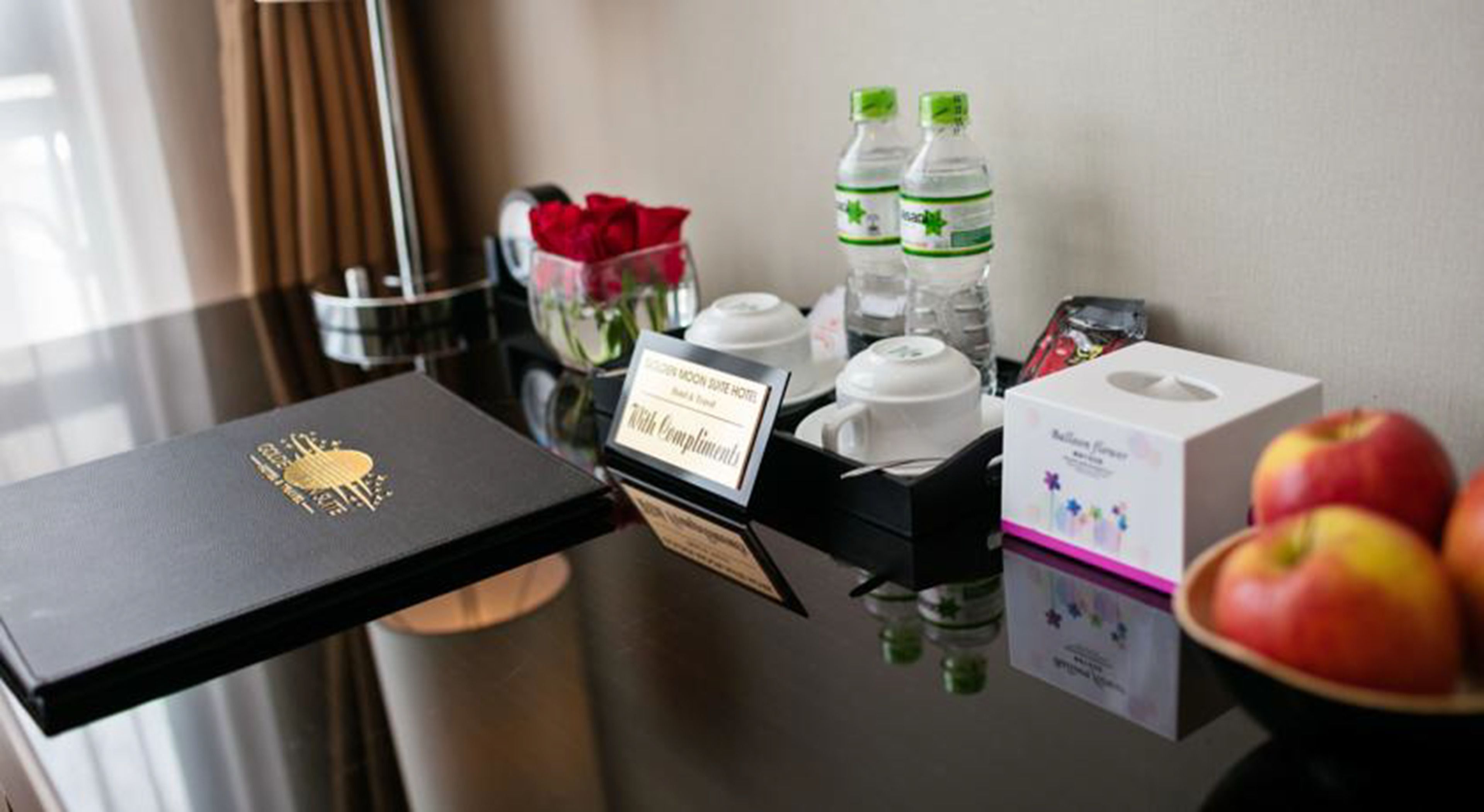 A hotel room with a black tray table, a gold emblem, a "Thank You Compliments" sign, two water bottles, a tissue box, and a bowl of apples.