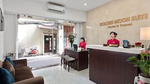 A reception area with a reception desk, a receptionist, and a sign that reads "Golden Moon Suite Hotel & Travel." There are two chairs and a couch in the foreground, and a bicycle is visible outside the entrance. - Golden Moon Suite Hotel (Hanoi)